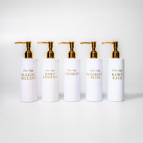 Fünf weiße Seifenspender aus der 'Pure Katy'-Kollektion: 'Blueberry Bliss', 'Magic Melon', 'Baby Banana', 'X-MAS Cookies' und 'Kiwi Kiss', mit goldenen Pumpen auf einem rosa Hintergrund mit Schaumblasen.